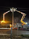 Utility trucks with raised buckets repair a power pole at night along a rural road.