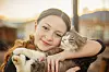 Smiling girl holding a fluffy gray and white cat outdoors at sunset.